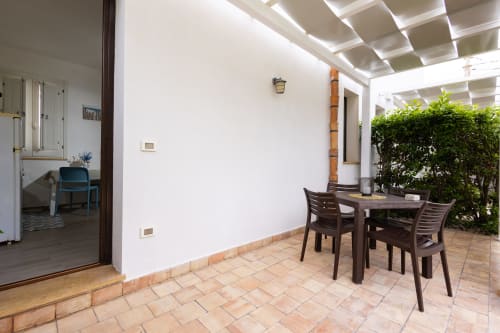 Covered terrace with dining table and chairs, white walls, green plants, and interior room view at Senia del Rais, Favignana