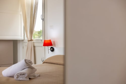 Bright bedroom at Senia del Rais in Favignana with red lamp, window, towels on bed, and ocean view curtains