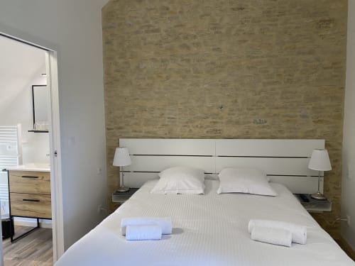 Modern studio bedroom at Domaine des Piéris with white bedding, cork accent wall, bedside lamps, and en-suite bathroom