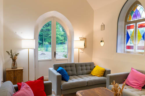 Élégant salon de style chapelle au Château De La Malmaison avec canapé gris, coussins colorés, fenêtres cintrées et vitraux