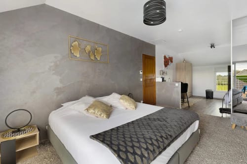 Chambre moderne avec vue sur jardin au Gîte Le Petit Villeneuve, lit blanc, mur gris, coussins dorés et
