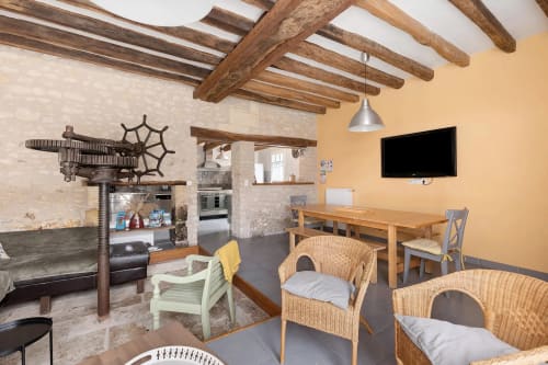 Salon rustique du gîte Le Petit Villeneuve avec poutres apparentes, murs en pierre, chaises en osier et télévision