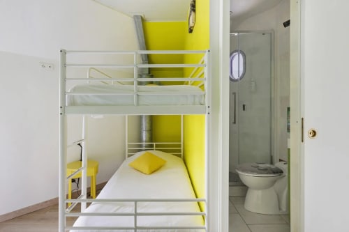 Lit superposé en métal blanc avec accents et coussins jaunes dans une chambre lumineuse, salle de bain attenante visible au Petit Villeneuve