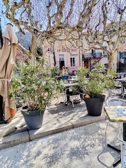 Freiluft-Café-Terrasse mit kahlen Platanen, Topfpflanzen und rosa Gebäude in Gagnières, Südfrankreich
