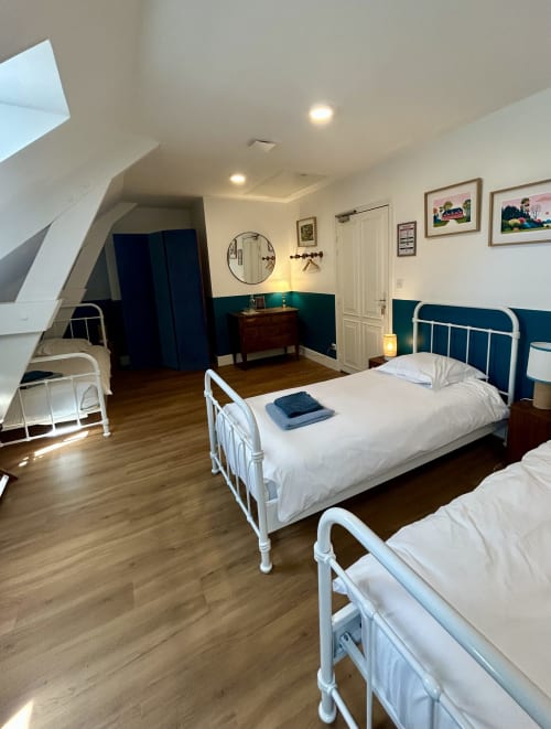 Chambre dortoir spacieuse au Manoir De Daubeuf avec lits en métal blanc, mur accent teal et puits de lumière.