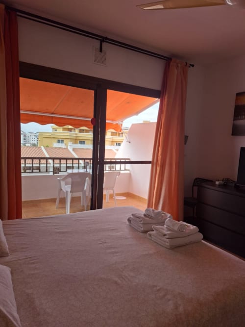 Bright bedroom with balcony overlooking Costa Adeje, orange awning, white chairs, folded towels, TV and dark furniture