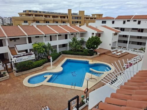 Aerial view of Costa Adeje resort pool with white loungers, terracotta roofs, and modern apartment buildings under cloudy sky