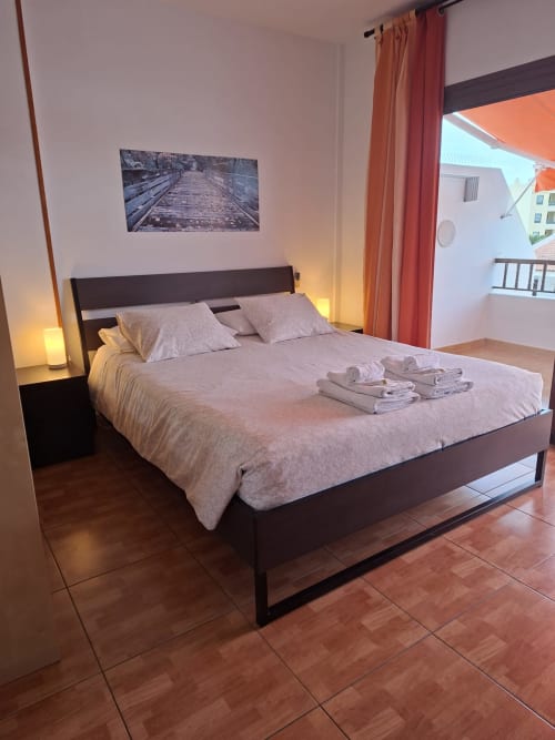 Modern bedroom in Costa Adeje apartment with dark bed, beige bedding, folded towels, wooden flooring, and ocean view balcony