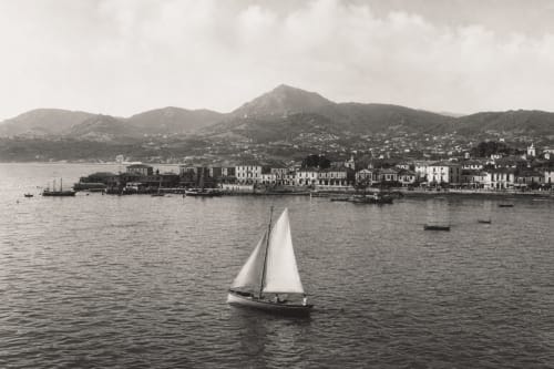 Velero en el agua frente al pueblo costero de Baiona con montañas, España