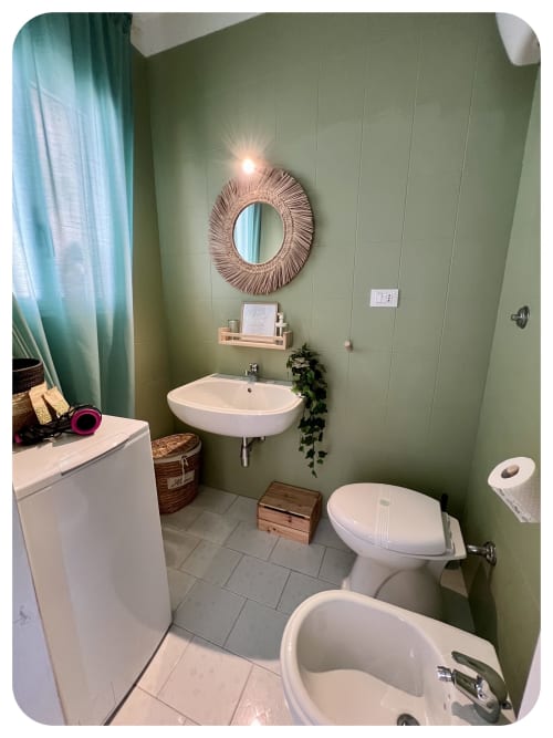 Sage green bathroom with wall-mounted sink, toilet, bidet, and woven circular mirror at Dimore Adua villa in Ostuni