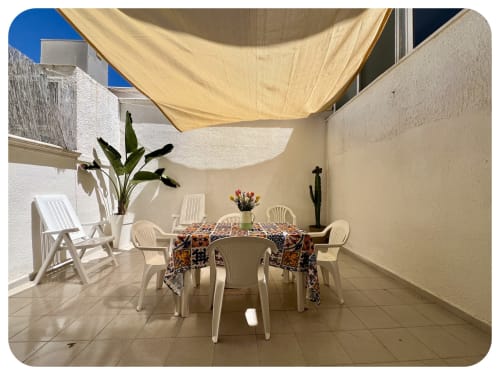 Sunny outdoor patio at Villa Adua in Ostuni with white furniture, dining table, shade sail, and garden plants