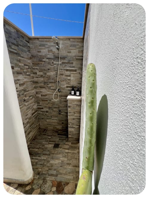 Outdoor shower at Dimore Adua villa in Ostuni with tiled walls, showerhead, and potted cactus plant against blue sky