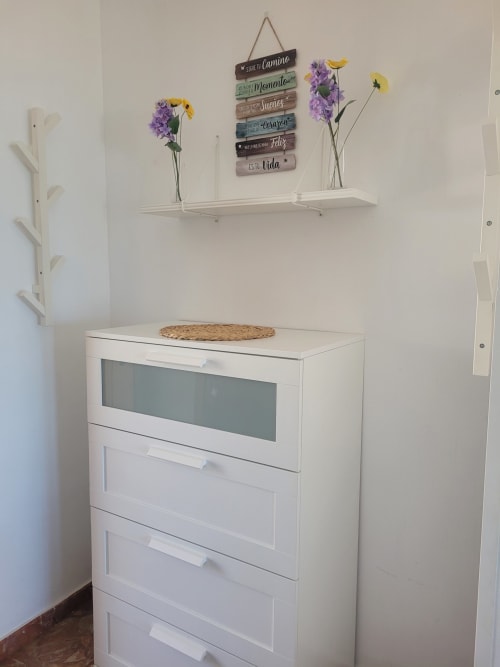 Unidad de almacenamiento blanca con estante decorativo, flores moradas y cartel de madera inspirador en primera línea de playa de Matalascañas