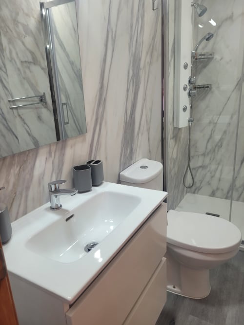Baño moderno en apartamento de Matalascañas con lavabo blanco, inodoro, azulejos efecto mármol y cabina de ducha