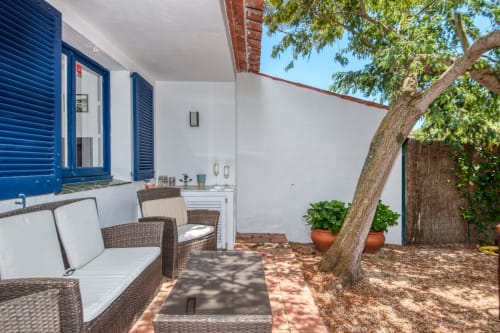 Patio d'hôtel à Palafrugell avec mobilier en rotin, volets bleus, pots en terre cuite et grand arbre offrant de l'ombre