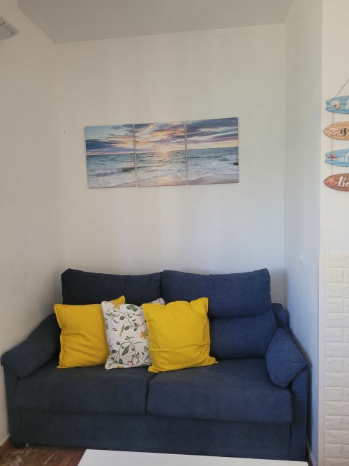 Sofá azul oscuro con cojines amarillos y cuadro de atardecer en playa en la sala de estar del apartamento de Matalascañas