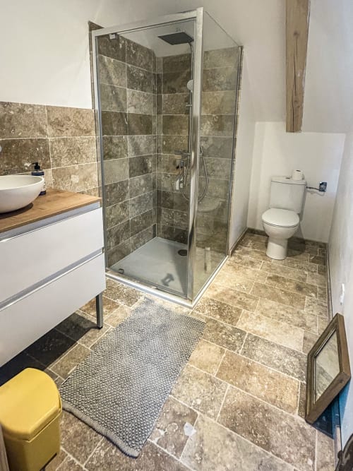 Salle de bain moderne à la Cour Du Liège avec douche ouverte, lavabo blanc, toilettes et carrelage en travertin