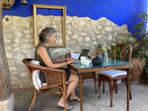 Femme travaillant sur son ordinateur dans la cour de Casa Gallinera à Alicante, murs en pierre rustique et pan bleu.