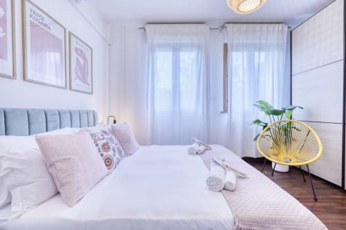Camera da letto moderna con testata blu, cuscini rosa, biancheria bianca e poltrona gialla con pianta in Luxury B&B a Pisa