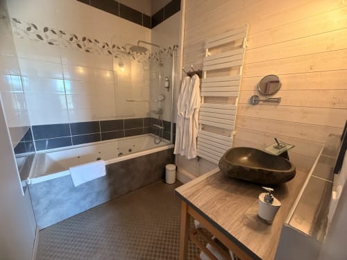 Salle de bain moderne avec baignoire balnéo, vasque en pierre noire, sèche-serviettes chauffant et touches de bois aux Instants Volés BnB