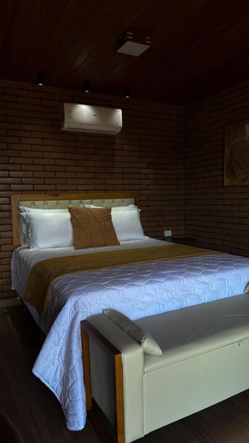 Quarto da villa Vivendas Canaã com cama queen, almofadas brancas, detalhes marrom, paredes de tijolos e ar condicionado de parede