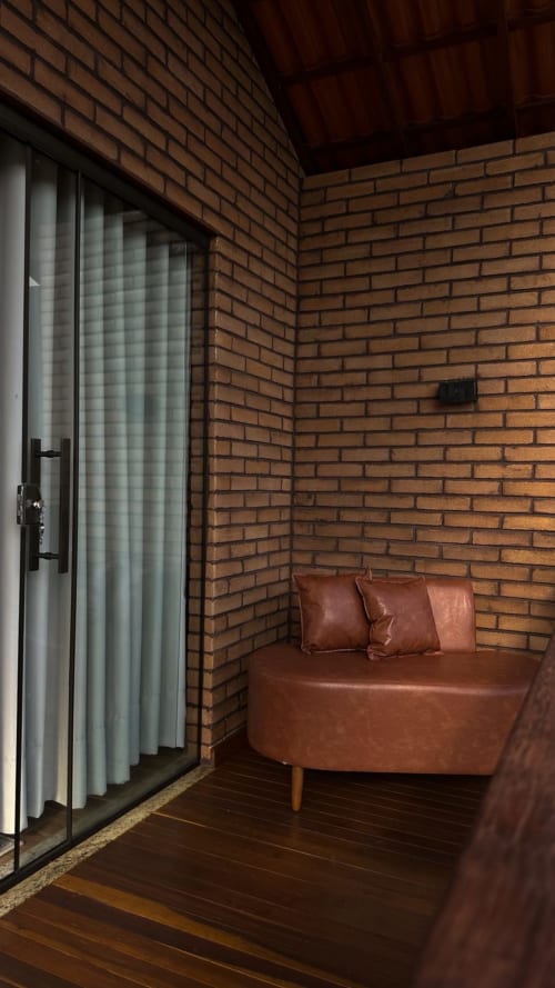 Interior aconchegante da villa Vivendas Canaã com sofá de couro marrom, paredes de tijolos, piso de madeira e porta de vidro com cortinas brancas