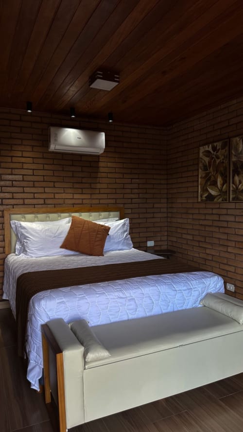 Quarto aconchegante com tijolos à vista, teto de madeira, cama climatizada com roupa branca, banqueta e ar-condicionado na parede