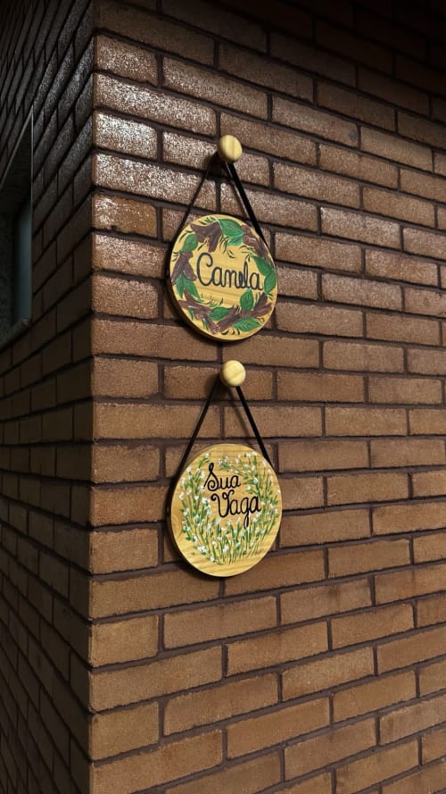 Duas placas decorativas de madeira penduradas em parede de tijolos em Vivendas Canaã: 'Camela' e 'Boa Vaga' com designs botânicos