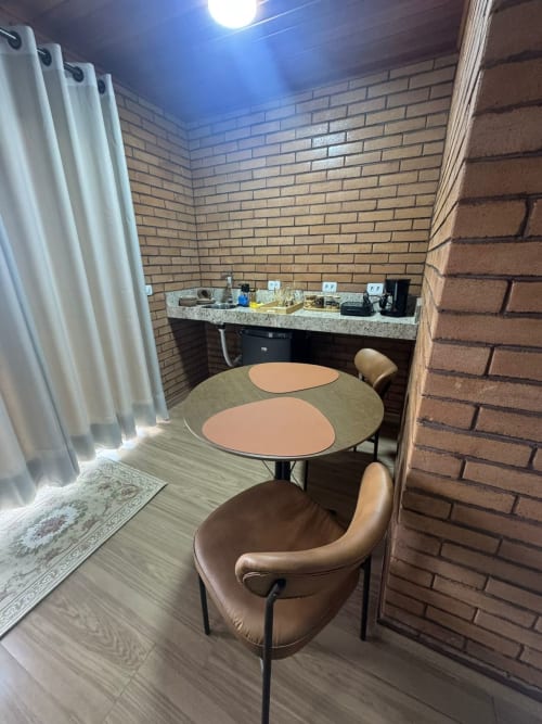 Sala de jantar do Chalé Canela com mesa redonda, cadeiras em couro, paredes de tijolos, kitchenette e cortinas brancas em Vivendas Canaã