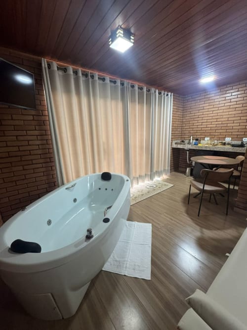 Banheiro spa luxuoso com banheira de hidroterapia branca, paredes de tijolos, cortinas elegantes e piso de madeira em Vivendas Canaã