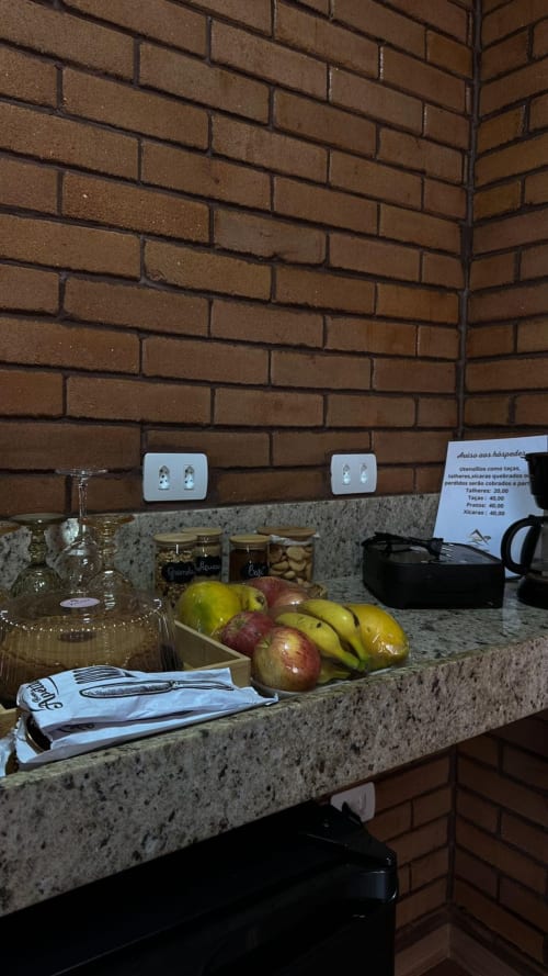 Bancada da cozinha em Vivendas Canaã com superfície em granito, frutas frescas, utensílios de vidro e tomadas brancas na parede de tijolos