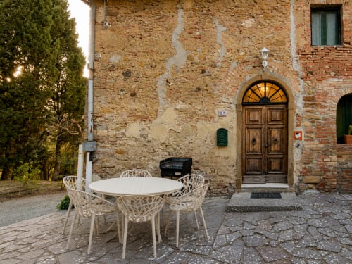 Tuscan stone villa courtyard with white dining chairs around table, historic wooden arched doorway, Lajatico Italy