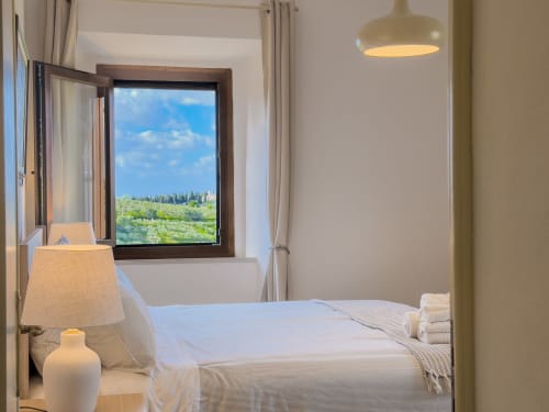 Bright Tuscan bedroom with mountain view, white bedding, pendant light, and open window overlooking Lajatico hills