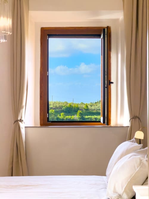 Serena apartment bedroom with open wooden window framing Tuscan hillside views, white bedding, and natural light