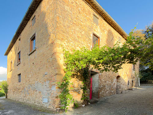 Golden stone Tuscan apartment building with ivy, red door, and clear blue sky in Lajatico