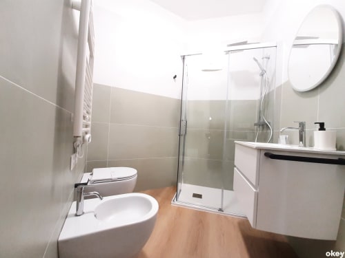 Bagno moderno nell'hotel di Bari con sanitari bianchi, doccia in cristallo, pavimentazione in legno e specchio rotondo