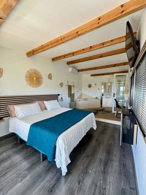 Chambre principale moderne avec lit queen-size, poutres en bois apparentes, plaid bleu, sol foncé et salle de bain attenante aux Villas d'A