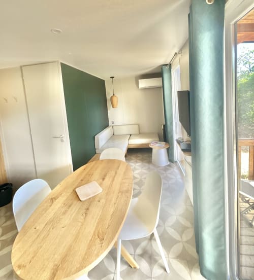 Espace de vie moderne du mobil-home avec table à manger en bois, mur vert menthe, canapé et vue sur le jardin au Domaine Pero Longo