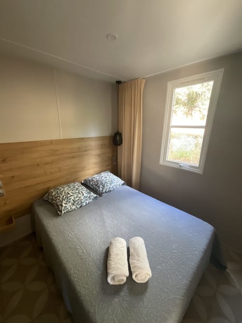 Chambre lumineuse du mobilhome Sartène avec tête de lit en bois, vue sur le jardin et deux serviettes roulées sur le lit