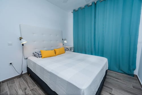 Dormitorio moderno en Smart Rent Fuerteventura con cama blanca, almohadas amarillas, cortinas turquesas y suelo de madera oscura