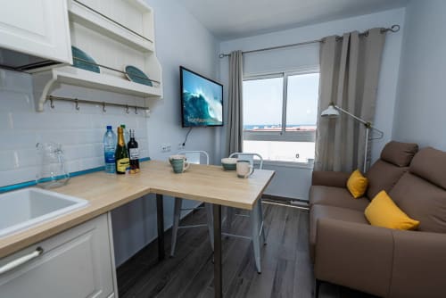 Moderno estudio en Tarajalejo, Fuerteventura con cocina equipada, sofá marrón con cojines amarillos, TV y vista al mar