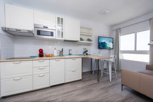 Cocina moderna blanca con encimera azul, electrodomésticos de acero inoxidable, barra y TV en apartamento Casa Sol, Tarajalejo