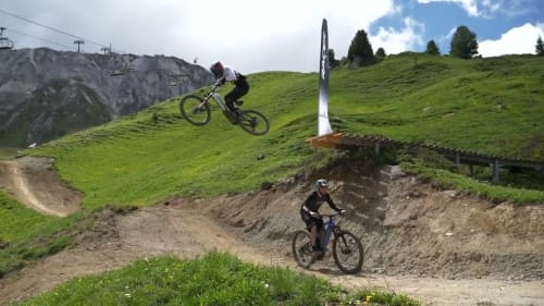 Vététistes effectuant des sauts sur les pistes du Chalet des Cimes, Belle Plagne, avec pentes vertes et remontées mécaniques visibles.