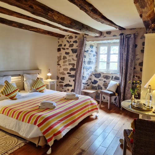 Chambre cosy au Mas De Rigoulac avec poutres apparentes, literie rayée, murs en pierre et vue sur le jardin à Aubrac