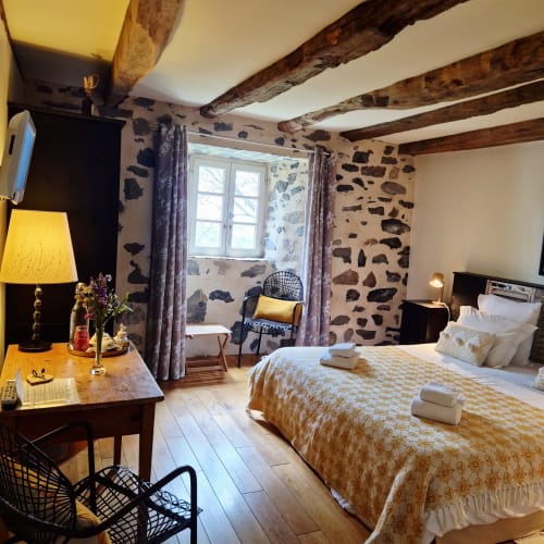 Chambre zen avec poutres apparentes, murs en pierre, lit queen-size avec parure jaune et vue sur le jardin au Mas De Rigoulac