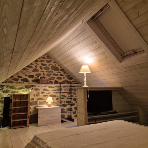 Chambre douillette sous les combles du Gîte Le Four avec murs en pierre, plafond en bois, TV, lampe et mobilier confortable