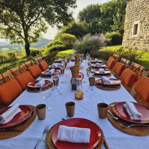 Élégante table dressée en extérieur au Mas De Rigoulac avec assiettes rouges, chaises oranges rembourrées et vue sur la campagne d'Aubrac