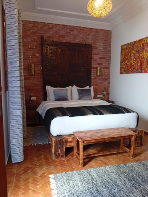 Chambre du Riad Le Village Mogador à Essaouira avec mur en brique rouge, tête de lit ornée sombre et banc traditionnel en bois