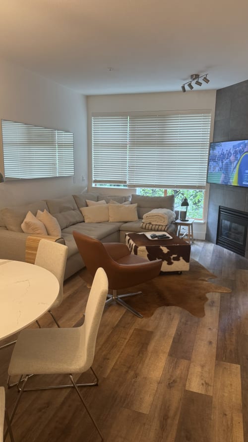 Modern Whistler apartment living room with sectional sofa, leather chair, fireplace, TV, and mountain views through blinds