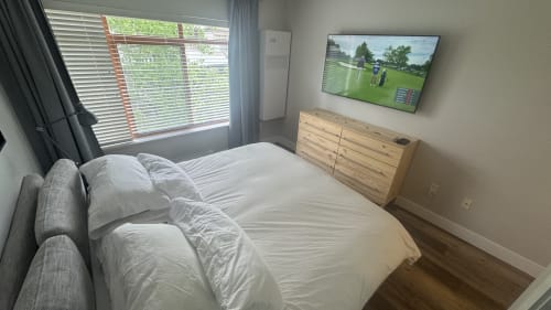 Modern bedroom in Whistler apartment with queen bed, wooden dresser, mounted TV showing golf, and mountain view through